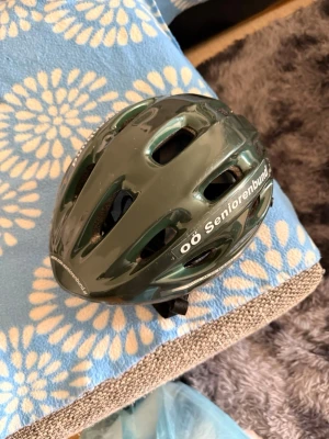 Grön cykelhjälm med text - Säljer en grön cykelhjälm med flera ventilationshål och texten 'oö Seniorenbund' på sidan. Hjälmen har justerbart spänne under hakan och är tillverkad i plast med skumvaddering på insidan.