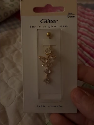 Glitter navelpercing med cubic zirconia - Snygg navelpercing från Glitter med en bar i kirurgiskt stål och cubic zirconia-stenar. percingen har en guldig färg och en elegant form med flera gnistrande stenar som bildar ett dekorativt mönster. Perfekt för dig som gillar glittriga accessoarer och vill sticka ut.