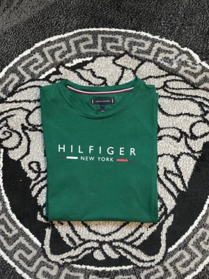 Grön Hilfiger New York t-shirt - Snygg grön t-shirt från Tommy Hilfiger med trycket 'HILFIGER NEW YORK' på bröstet. Klassisk passform med korta ärmar och rund hals. Tillverkad i mjuk bomull som känns skön mot huden. Perfekt för dig som gillar stilrena och ikoniska plagg.