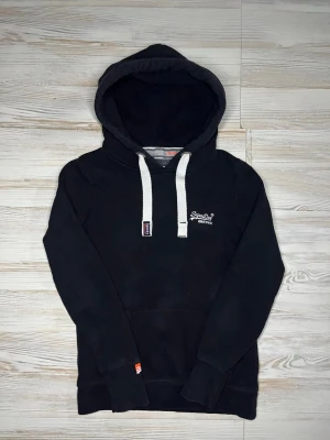 Svart hoodie från Superdry - Snygg svart hoodie från Superdry med vit broderad logga på bröstet🤩 Hoodien är i bra skick och passar till allt, Storlek:Xs🙌🏻 Hör av er vid snabbaffärer samt funderingar.😇