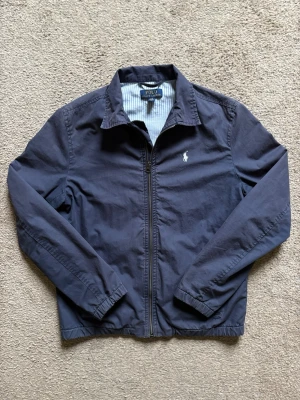 Marinblå jacka från Polo Ralph Lauren - Klassisk marinblå jacka från Polo Ralph Lauren med dragkedja framtill och broderad logga på bröstet. Jackan har krage, muddar och randigt innerfoder. Perfekt för dig som gillar stilren och tidlös design.
