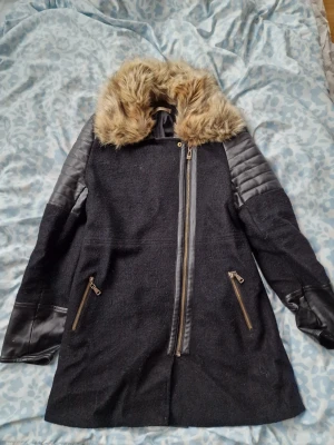 Svart kappa med fuskpäls från Vera & Lucy - Stilren svart kappa från Vera & Lucy med stora detaljer i fuskpäls runt kragen och quiltade skinnimitationer på ärmarna. Asymmetrisk dragkedja framtill och två snedställda fickor med dragkedja. Perfekt för dig som vill ha en edgy och trendig look. OBS hål i ena fickan som enkelt går att laga för den som orkar!