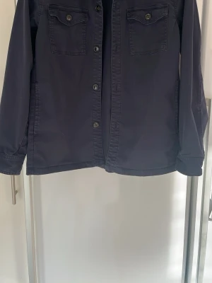 Mörkblå overshirt med fickor - Stilren mörkblå overshirt med knäppning framtill och två bröstfickor med lock och knapp. Långärmad modell i ett mjukt bomullsliknande material. Perfekt att bära som lager över t-shirt eller hoodie. I stortsätt oanvänd! Storlek 176, största storleken innan vuxenstorlekar.
