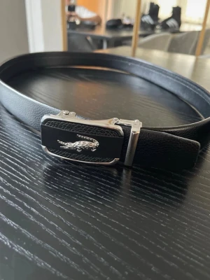 Svart skärp/bälte - Snyggt svart skärp  i skinn med silverfärgat krokodilemblem på spännet. Bältet har en stilren design och metallspänne med detaljer i svart och silver. Perfekt för att lyfta vilken outfit som helst.