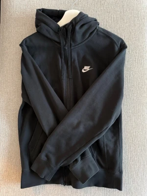 Svart Nike hoodie med dragkedja - Snygg svart hoodie från Nike med dragkedja framtill och vit Nike-logga på bröstet. Hoodien har huva med snörning och två fickor på sidorna. Tillverkad i mjukt och bekvämt material, perfekt för en avslappnad stil.
