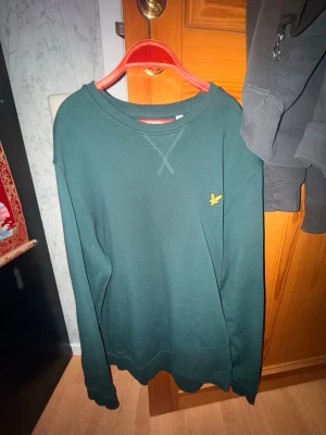 Mörkgrön sweatshirt från Lyle & Scott - Mörkgrön sweatshirt från Lyle & Scott i ekologisk bomull. Klassisk modell med rund halsringning, ribbade muddar och den ikoniska gula logon broderad på bröstet. Enkel och stilren design som funkar till det mesta. Perfekt basplagg för garderoben.