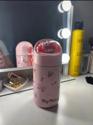 My Melody rosa termosflaska - Söt rosa termosflaska med My Melody-motiv från Sanrio. Flaskan har små tryck av jordgubbar, rosetter och hjärtan samt ett genomskinligt lock med en My Melody-figur inuti. Praktisk silikonögla baktill för enkel bärning. Perfekt för drycker på språng.