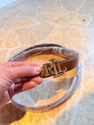 Brunt skärp från Ralph Lauren - Snyggt brunt skärp från Ralph Lauren med ett stort guldfärgat RL-spänne framtill. Bältet är gjort i skinn och har en klassisk, stilren design som passar till många olika outfits. Perfekt accessoar för att lyfta din look.