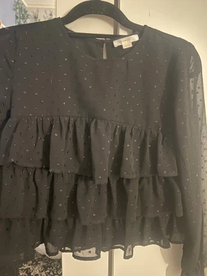 Svart volangblus med prickar - Trendig svart blus från Gina Tricot med små silvriga prickar och flera lager volanger framtill. Blusen har rund halsringning och är tillverkad i ett tunt, transparent material som ger en luftig känsla. Perfekt för dig som gillar en lekfull och chic stil.