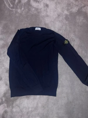 Mörkblå stickad tröja Stone Island - Säljer en mörkblå stickad tröja från Stone Island med den klassiska logotypen på vänster ärm. Tröjan har rund halsringning, ribbade muddar och är tillverkad i ett mjukt stickat material. Perfekt för dig som gillar streetwear och snygga detaljer.