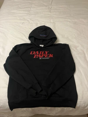 Svart hoodie från Daily Paper - Svart hoodie från Daily Paper med röd text 'DAILY PAPER BE VIGILANT' på bröstet och broderad logga på huvan. Klassisk passform med känguruficka och ribbade muddar. Perfekt för en chill och stilren look. Riktig kvalitetshoddie