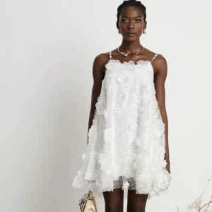 Vit blommig kortklänning med pärlband - Superfin vit kortklänning med 3D-blommor, pärlprydda axelband och glittriga detaljer. Klänningen är helt ny då jag skulle ha den till min student men den kom aldrig i tid, den är ifrån miss-rosier 💕 passar. XS-S