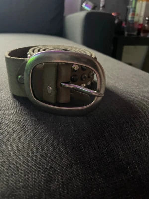 Brunt skärp med nitar och metallspänne - Coolt brunt skärp med silverfärgade nitar och ett robust metallspänne. Skärpet är gjort av skinn och har ett klassiskt hålmönster längs hela bandet. Perfekt för att ge din outfit en edgy touch.