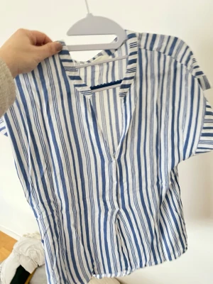 Blåvit randig skjorta från Stories - Kortärmad skjorta från Stories i blått och vitt randigt mönster. Skjortan har v-ringning, knappar framtill och är tillverkad i ett lätt och luftigt material, perfekt för varmare dagar. Passar dig som gillar en avslappnad men ändå stilren look.