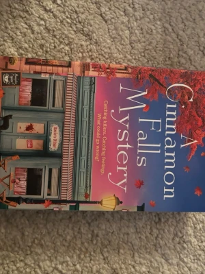A Cinnamon Falls Mystery bok - En färgstark pocketbok med titeln 'A Cinnamon Falls Mystery'. Omslaget har en mysig småstadskänsla med röda höstlöv, blå och rosa toner samt en butiksmiljö. Perfekt för dig som gillar mysterier och småstadsliv.