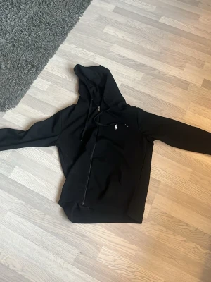 Svart hoodie från sm Polo Ralph Lauren - Snygg svart hoodie från Polo Ralph Lauren med dragkedja och huva. Klassisk logga broderad på bröstet. Tillverkad i mjukt material som känns skönt mot huden. Perfekt för en avslappnad och stilren look.