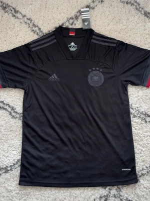 Svart Tyskland matchtröja Adidas - Snygg svart matchtröja från Adidas med Tysklands landslagsmärke och fyra stjärnor på bröstet. Tröjan har korta ärmar, V-ringning och diskreta grå ränder på axlarna. Tillverkad i lätt och ventilerande material med Aeroready-teknologi.