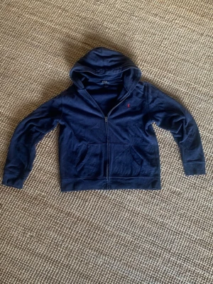 navy blå Polo Ralph Lauren zip hoodie - Mörkblå hoodie med dragkedja från Polo Ralph Lauren. Klassisk design med huva, kängurufickor och rött broderat logotyp på bröstet. Mjuk bomullsblandning som är skön att ha på sig. jag vill bli av med den pris ej hugget i sten. den är ganska sliten men det är inte så stora defekter, Inga hål. Från axel till midjan är måttet 60cm (se bild 4). Från axel till hand är måttet 60cm (se bild 5). Den är i storlek XL för barn, men den är ganska kort ( kontrollera måtten). DM om ni har några funderingar.