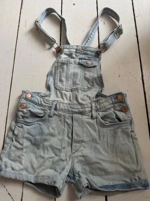Ljusblå jeansshorts med hängslen - Säljer ett par ljusblå jeansshorts med hängslen och bröstficka. Shortsen har klassiska jeansknappar i kopparfärg, fickor fram och bak samt justerbara axelband. Perfekta för varma dagar och en avslappnad stil.