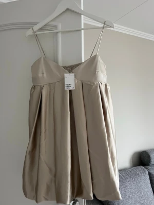 Beige kort klänning med tunna band - Säljer en beige kort klänning med tunna axelband och puffig kjol. Klänningen har en söt ballongformad siluett och smock i ryggen för extra komfort. Perfekt för sommarens alla tillfällen och ger en trendig look.