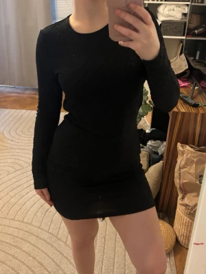 Svart glittrig kort festklänning - Snygg svart kort klänning med diskret glittrigt mönster. Klänningen har lång ärm och en tight passform som framhäver figuren. Perfekt för dig som vill sticka ut med en stilren look.