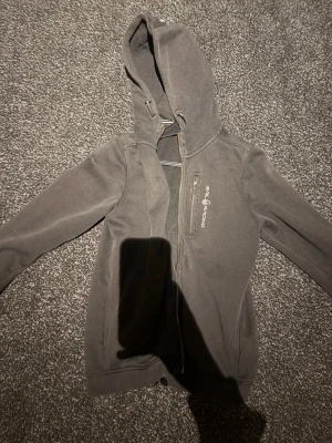 Mörkgrå hoodie från Sail Racing - Mörkgrå hoodie från Sail Racing med dragkedja framtill och justerbar huva. Tillverkad i mjukt fleecematerial som känns skönt mot huden. Diskret logga på bröstet och två fickor på sidorna. Perfekt för chill dagar eller när du vill ha något bekvämt. Priset är som vanligt förhandlingsbart.