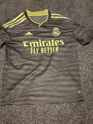 Real Madrid bortatröja Modric #10 - Snygg svart bortatröja från Real Madrid med gula detaljer och tryck. Adidas-logga, klubbmärke och sponsortryck på framsidan samt Modric och nummer 10 på ryggen. Tillverkad i lätt och ventilerande material, perfekt för fotbollsträning eller match. Väldigt bra skick bortsett ifrån lite släpp av bokstäverna. Priset är förhandlingsbart