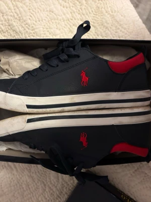 sneakers från Polo Ralph Lauren - Mörkblå sneakers från Polo Ralph Lauren Scranton med röd logga på jackan och röd detalj vid hälen. Använts max 3 gånger och säljs då de tyvärr är lite små