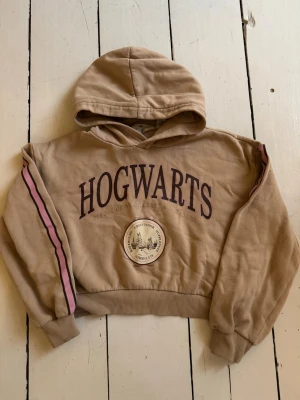 Beige Hogwarts hoodie med tryck - Cool beige hoodie med stor HOGWARTS-text och emblem framtill. Har huva och ribbade muddar, samt rosa och lila ränder längs ärmarna. Kortare tröjmodell. Perfekt för dig som älskar Harry Potter och vill ha en avslappnad stil.