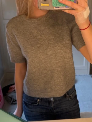 Grå stickad kortärmad tröja - Mysig grå stickad tröja med korta ärmar och rund halsringning. Tröjan har en enkel och stilren design, perfekt att matcha med jeans eller kjol. Materialet ser ut att vara mjukt och lite fluffigt, vilket ger en avslappnad vibe.