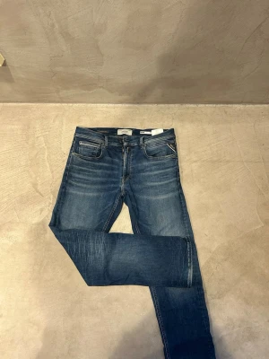 Blå slim fit jeans från Replay - Snygga blå slim fit jeans från Replay med klassisk femficksdesign och diskreta slitningar framtill. Jeansen har en brun patch med Replay Hyperflex-logga bak i midjan och kontrastsömmar på bakfickorna. Perfekta för en avslappnad och trendig look.