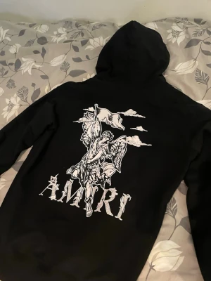 Svart hoodie från Amiri med tryck - Säljer en svart hoodie från Amiri med stort grafiskt tryck på ryggen som föreställer en ängel med flagga och texten 'AMIRI'. På bröstet finns ett mindre tryck med samma tema. Klassisk huva och känguruficka framtill. Perfekt för dig som gillar streetwear och unika prints.