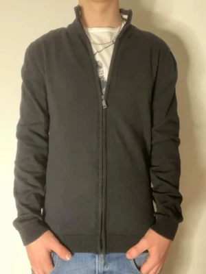 Svart ziptröja från McNeal - Snygg svart ziptröja från McNeal i ekologisk bomull. Tröjan har hög krage, dragkedja framtill och ribbade muddar vid ärmslut och nederkant. Perfekt till jeans eller över en t-shirt för en clean look.