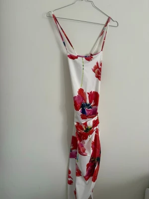 Vit blommig långklänning - Säljer en vit långklänning med tunna axelband och stora röda blommor. Kläningen sitter tajt på kroppen och har en fin öppen rygg 