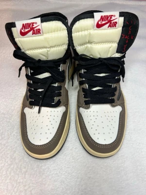 Nike Air Jordan 1 Travis Scott sneakers - Säljer ett par Nike Air Jordan 1 Travis Scott sneakers med bruna, beige, vita och svarta paneler. Skorna har svart snörning, ikonisk omvänd swoosh och broderad logga på hälen. Ovandelen är i skinn och mocka med röda Nike Air-detaljer på plösen. Skorna är OANVÄNDA.
