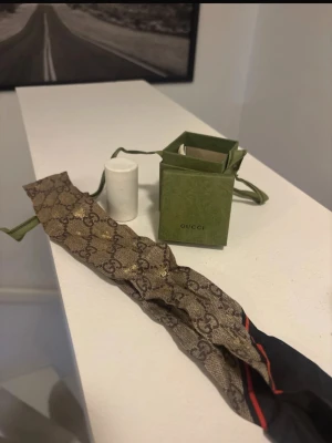 Gucci sjal med monogram och röd detalj - Säljer en exklusiv sjal från Gucci med klassiskt GG-monogram i beige och brunt. Sjalen har en smal, avlång form och en snygg röd detalj i kanten. Materialet är troligtvis silke eller bomull, och den levereras med originalförpackning. Perfekt för dig som vill ha något lyxigt och trendigt.