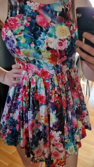 Färgglad blommig klänning från Ginger Fizz - Superfin ärmlös klänning med färgstarkt blommönster i rosa, blått, rött och vitt. Modellen är kort med rund halsringning och intressant rygg. Perfekt för sommarens alla tillfällen!