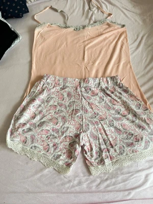 Ljusrosa pyjamas-set med spetsdetaljer - Sött pyjamas-set med ett ljusrosa linne med tunna axelband och spetskant samt matchande shorts i vitt med paisleymönster i rosa och grått. Shortsen har elastisk midja och spetskant nedtill. Perfekt för mysiga kvällar och nätter.