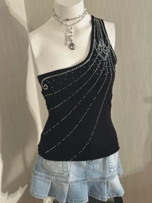 Svart oneshoulder topp med pärlor - Snygg svart oneshoulder topp med silverfärgade pärlor och detaljer som ger en glittrig effekt. Toppen har en unik asymmetrisk design och sitter tight mot kroppen. Perfekt för dig som vill sticka ut med coola detaljer.