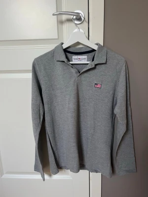 Grå långärmad skjorta från Marqy Classic - Snygg grå långärmad skjorta från Marqy Classic med krage och knappar framtill. Skjortan har en liten amerikansk flagga broderad på bröstet och är tillverkad i mjuk bomull. Perfekt för en clean och stilren look.