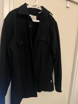 Svart overshirt med bröstfickor från Jack&jones  - Stilren svart overshirt från Jack&jones med klassisk krage och två bröstfickor med knappar. Overshirten har raka ärmar och knäppning framtill. Perfekt att bära som lager över t-shirt eller hoodie. Materialet är kraftigt och ger en snygg struktur.