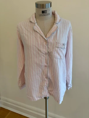 Rosa randig pyjamasskjorta Victoria's Secret - Söt pyjamasskjorta från Victoria's Secret i ljusrosa och vita ränder. Skjortan har långa ärmar, klassisk krage, knappstängning framtill och en bröstficka. Tillverkad i mjukt bomullsmaterial för extra komfort.