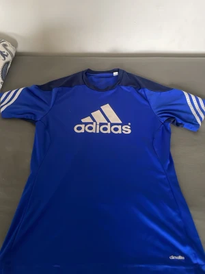 Blå Adidas t-shirt med vita detaljer - Säljer en blå sportig t-shirt från Adidas med vita logotypen på bröstet och vita ränder på ärmarna. Materialet är lätt och andas, perfekt för träning eller sport. Climalite-tyget håller dig torr och bekväm under aktivitet.