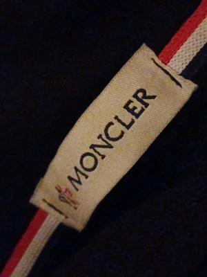 Svart jacka moncler - Snygg svart hoodie från Moncler med huva och klassiska Moncler-detaljer. Tröjan har ribbade muddar med rödvita ränder och en enkel, stilren design. Perfekt för dig som vill ha en exklusiv och bekväm hoodie med streetwear-vibe.