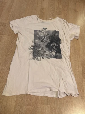 Vit t-shirt med blommigt tryck - Vit t-shirt med svartvitt blommigt tryck och texten 'WILDFLOWER' på framsidan. Klassisk rund hals och korta ärmar. Perfekt för en avslappnad stil och lätt att matcha med jeans eller shorts.
