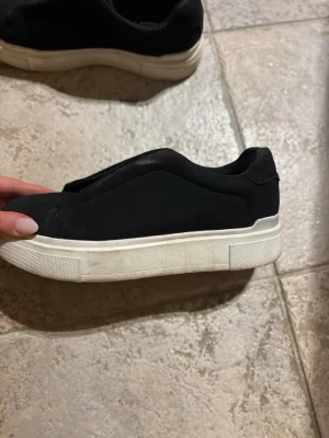 Svarta slip-on sneakers från 157 - Stilrena svarta slip-on sneakers från 157 med tjock vit sula. Ovandelen är i textil och skon har en minimalistisk design utan snörning. Perfekta för dig som gillar enkel och clean stil. Använda men går att göra rent egentligen bara smutsiga. Fint skick annars inget trasigt