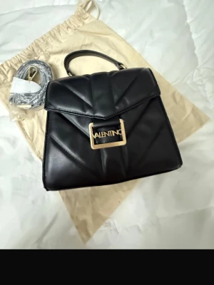 Svart handväska från Valentino - Snygg svart handväska från Valentino med quiltad design och guldfärgat metallspänne med logotyp. Väskan har ett topphandtag och medföljande axelrem. Tillverkad i konstläder och har en elegant, strukturerad form. Perfekt accessoar för att lyfta din outfit.