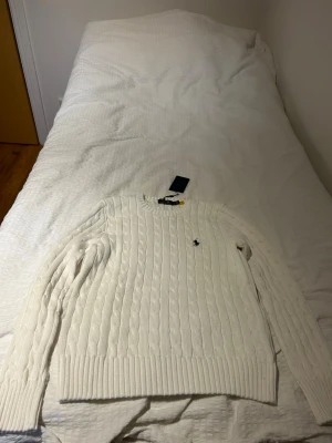 Vit kabelstickad tröja Polo Ralph Lauren - Kabelstickad tröja i offwhite från Polo Ralph Lauren med rund halsringning och diskret broderad logga på bröstet. Tröjan har långa ärmar och breda ribbade muddar vid ärmslut och nederkant. Klassisk och tidlös design i mjukt material.