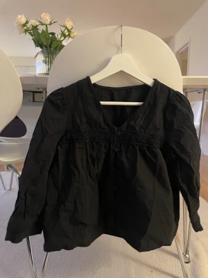 Svart blus med spetsdetaljer - Snygg svart blus med v-ringning och dekorativa spetsdetaljer längs halsringningen och bröstet. Blusen har en lös passform och långa ärmar med lätt puff vid axlarna. Perfekt att styla med jeans eller kjol för en trendig look.