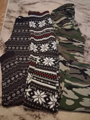 3-pack leggings med mönster - Säljer ett 3-pack leggings med olika mönster: ett par svarta med vita snöflingor och zigzag-mönster, ett par med juligt rött, vitt och svart snöflingemönster samt ett par i klassiskt grönt kamouflage. Perfekta för dig som vill ha både mysiga och coola stilar.